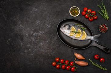 Redy to cook fresh dorado fish or sea bream on cast-iron pan with ingredients  lemon herbs cherry tomatoes and spices on dark background with copy space. Top view space for text.