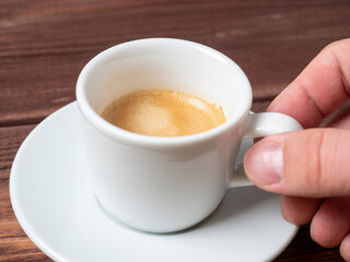 close-up of a man's hand holding a small white cup of aromatic espresso over a white saucer. Wooden background, side view.