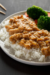 Homemade Orange Chicken with White Rice, Broccoli and Carrot on a plate on a black background, side view.