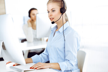 Blond woman call operator is using computer and headset for consulting clients online. Group of...
