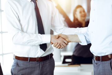 Fototapeta premium Two businessmen shaking hands in sunny office, close-up. Happy and excited business woman stands with raising hands at the background. Business people concept