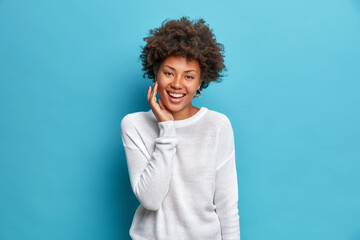 Happy dark skinned charmming woman with curly hair smiles broadly touches face enjoys soft skin has natural beauty expresses positive emotions dressed incasual jumper isolated on blue studio wall