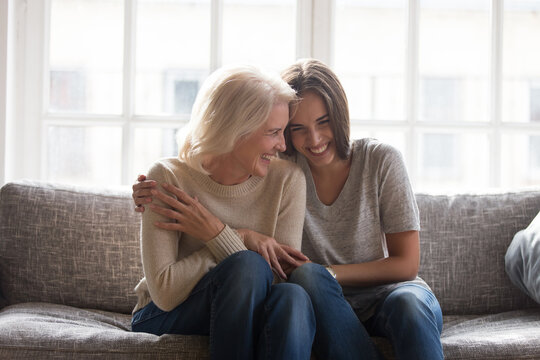 Smiling adult daughter and mature 60s mother sit relax on sofa at home have fun laugh enjoy family time together. Happy millennial grownup woman child cuddle hug with old mom on couch, show love care.