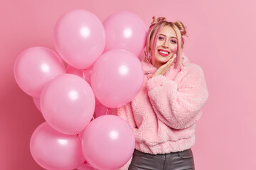 Happy young European woman poses with many balloons enjoys party on her birthday smiles gladfully wears fur coat has food festive mood awaits for start of holiday isolated over pink background