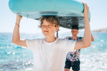 Blonde teenage son with father surfers smiling carrying the stand-up paddleboard with a paddle on...