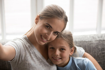 Smiling young Caucasian mother and small teen daughter make self-portrait picture on smartphone. Happy mom and little girl child have fun pose for family selfie on cellphone. Technology concept.