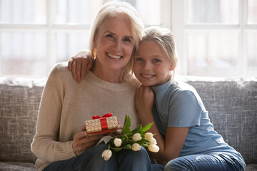 Portrait of happy elderly Caucasian grandmother and little teen granddaughter pose at home...
