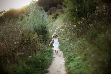A young blonde girl in a white dress runs along the path in the park barefoot, happy free childhood, closeness to nature, natural and careful upbringing