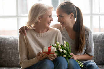 Happy caring young female congratulate greet overjoyed older Caucasian mother with birthday anniversary at home. Smiling loving adult grownup daughter make surprise present gift and flowers to mom.