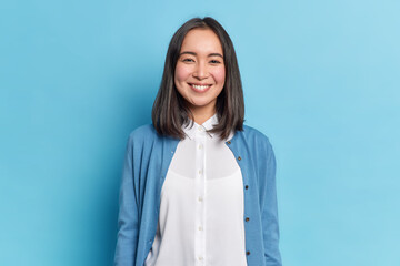 Horizontal shot of pretty Asian woman with dark hair smiles pleasantly looks directly at camera has toothy smile wears white shirt and jumper isolated over blue background. Emotions concept. - Powered by Adobe