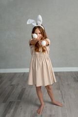 Funny smiling child having fun on Easter egg hunt. School girl ten years old wearing bunny ears on gray background. mischievous kid gives, holds out white egg and grinning. Holidays, spring, Easter co