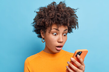 Emotional surprised African American woman stares at smartphone screen reads amazing news with surprised face being stunned by something wears casual orange jumper isolated over blue background. - obrazy, fototapety, plakaty