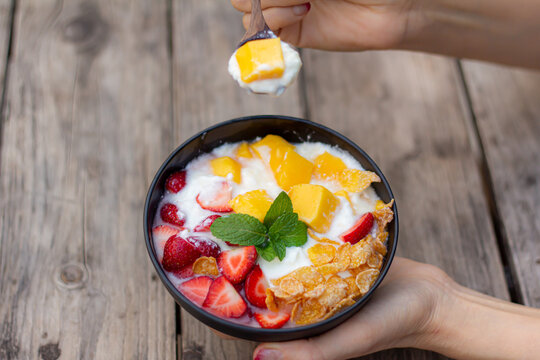 hand use a wood spoon to scoop greek yogurt homemade with mixed strawberries mango conflex mint topping in black bowl on wooden table closeup view