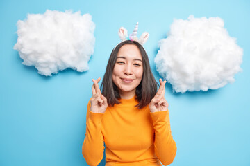 Good looking Asian woman with satisfied expression crosses fingers believes in good luck before important event wears unicorn headband orange jumper isolated over blue background. Body language