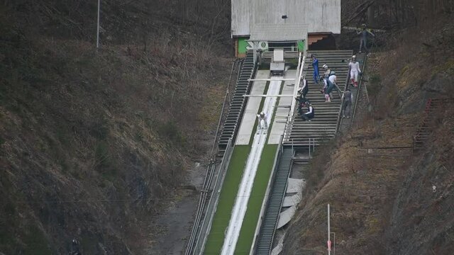 Ski jump ramp. Jumper runs down to generate sufficient speed before reaching the jump. Person glide down on tracks along the in-run. Static shot, slow motion