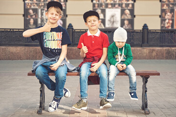 Obraz na płótnie Canvas Positive elder brothers in stylish caps show thumbs-up sitting near tired little boy on bench on contemporary city street
