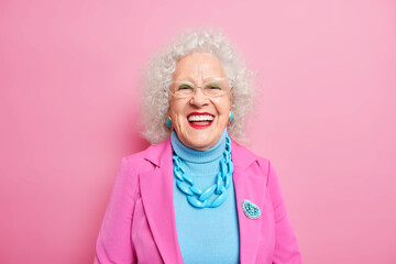 Portrait of aged beautiful woman with curly grey hair bright makeup smiles happily expresses positive emotions dressed in fashionable outfit isolated over pink background. Positive grandmother