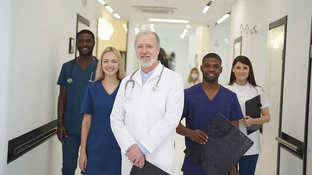 Portrait of senior doctor in white uniform and young doctors therapists posing looking at camera, multidisciplinary medical team in white coats in aisle