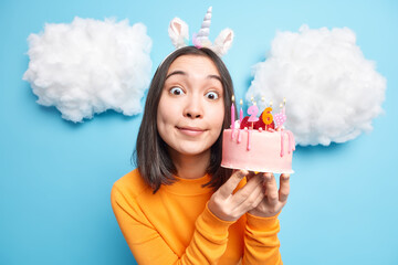 Lovely surprised brunette young Asian woman looks bugged eyes at camera wears casual clothes holds delicious cake isolated over blue studio background with white clouds overhead. Birthday concept
