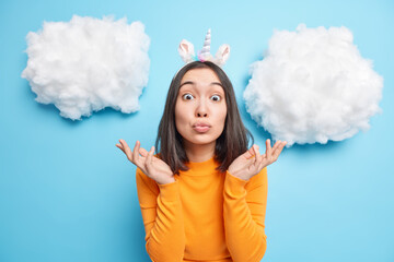 Surprised adorable Asian woman spreads palms keeps lips folded looks with shocked expression wears orange jumper unicorn headband has confused look isolated over blue background clouds above