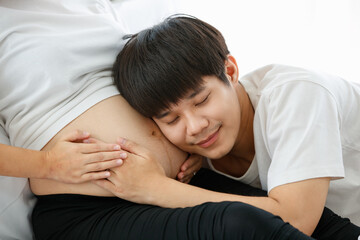 portrait of a very happy Asian man who will become to be a father. He is sitting and listening to a sound of a fetus in his pregnant wife's belly with love and happiness