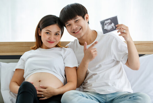 happiness young Asian couple wearing a casual dress smiling and sitting on a bed together. A happy husband puts his arm around his pregnant wife and showing an ultrasound scan photo of a fetus