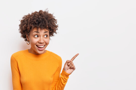 Horizontal Shot Of Positive Dark Skinned Woman Looks With Interest And Amazement Aside Indicates At Copy Space Shows Good Deal Recommends Product Wears Orange Jumper Isolated On White Studio Wall