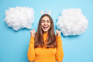 Horizontal shot of attractive happy brunette woman raises fists up exclaims from joy feels like winner enjoys luck dressed in orange jumper isolated over blue background white fluffy clouds above