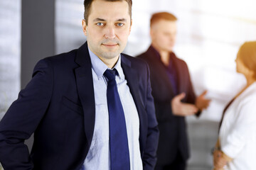 Fototapeta premium Portrait of a self-confident middle aged businessman in a blue suit, standing in a sunny modern office with his colleagues at the background. Concept of business success