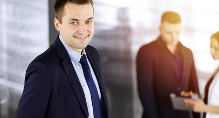 Portrait of a self-confident middle aged businessman in a blue suit, standing in a sunny modern...