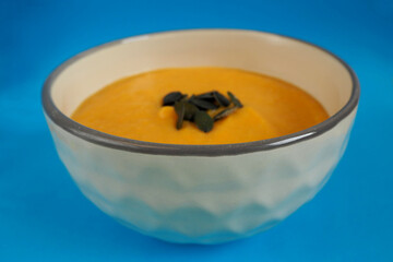 cream soup of pumpkin and carrot with pumpkin seeds stands in a round gray plate on a blue background side view . gluten-free homemade food