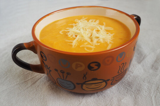 pumpkin and carrot cream soup with grated cheese is placed in a round brown plate with handles on the background of a beige linen tablecloth side view . homemade food