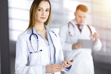 Professional woman doctor with a stethoscope is using a computer tablet, while she is standing together with her colleague in a sunny clinic. Young doctors at work in a hospital. Medicine concept