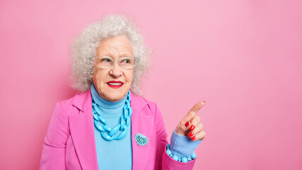 Mature beautiful woman with wrinkled skin wears stylish clothes jewelry indicates at upper right corner give wide advice shows copy space against pink backgroud. People age advertisement concept