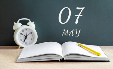 may 07. 07th day of the month, calendar date. White alarm clock, an open notebook with blank pages, and a yellow pencil lie on the table.Spring month, day of the year concept