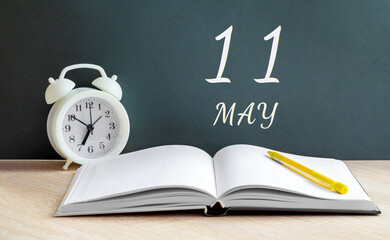 may 11. 11th day of the month, calendar date. White alarm clock, an open notebook with blank pages, and a yellow pencil lie on the table.Spring month, day of the year concept