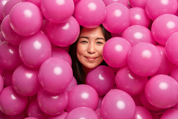 Fototapeta na wymiar Pleased young brunette Asian woman being happy at birthday party surrounded with pink inflated balloons smiles gently enjoys making photos near decor. People celebration and festivity concept