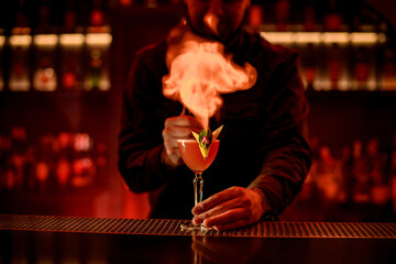 bartender sprinkles and sets fire over glass cocktail on bar counter. Red illumination.