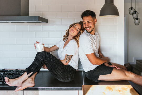 Happy Couple Sitting At The Kitchen Counter. Beautiful Woman Leaning On Her Handsome Man Who Is Sitting On Kitchen Counter, Both Are Smiling. Family Life 