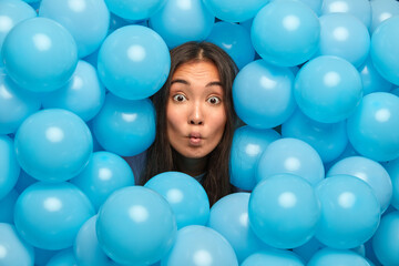 Fototapeta na wymiar Surprised brunette woman makes fish lips looks with funny grimace at camera has long dark hair sticks out head through inflated balloons awaits for festive holiday spends free time on party.