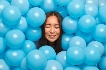 Fototapeta na wymiar People holiday decoration and celebration concept. Satisfied brunette Asian woman stands with closed eyes sticks out head through blue inflated balloons has delighted look dreams about something