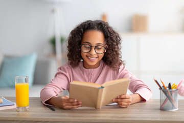 Leisure concept. Cute black schoolgirl reading paper book and drinking orange juice, sitting at desk