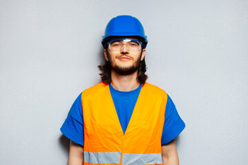 Studio portrait of young construction worker engineer wearing safety equipment; blue hard hat, transparent goggles and orange vest on the background of grey textured wall.