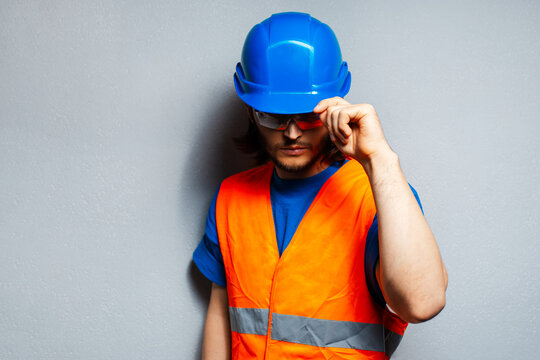 Studio portrait of young construction worker wearing safety equipment; blue hard hat, transparent goggles and orange vest on the background of grey wall.