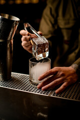 male bartender holds large piece of ice with tongs over steaming old-fashioned glass