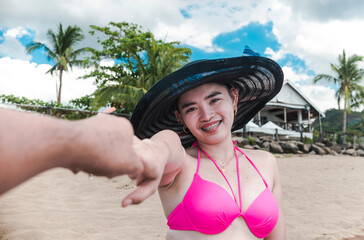A pretty asian woman in a large dark sun hat and pink bikini holds the hand of her boyfriend.