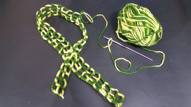 A piece of crocheted work in progress and a green and yellow ball of crochet wool or yarn. A purple needle is also in the shot.  On a black surface. 