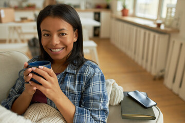 Happy young black woman in checkered shirt sitting on sofa with mug in her hands, drinking warm...