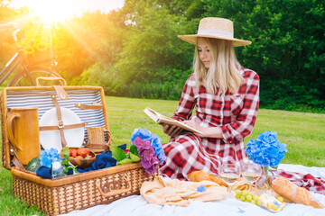 Girl in red checkered dress and hat sitting on white knit picnic blanket reading book and drinking wine. Summer picnic on sunny day with bread, fruit, bouquet hydrangea flowers. Selective focus