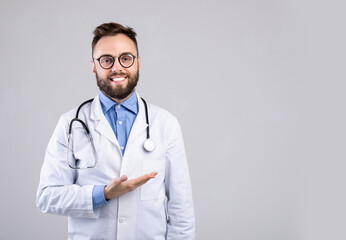 Cheerful male doctor in uniform pointing at empty space over grey studio background, panorama